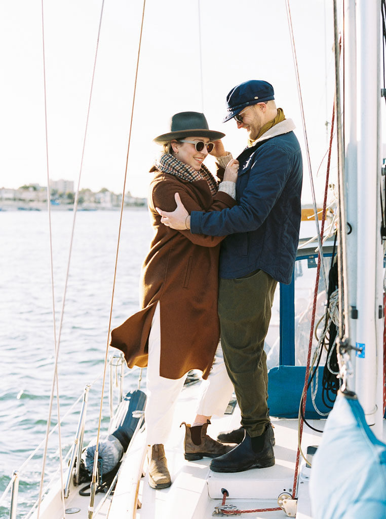 A woman is embraced by her finace as she hold onto her had and looks down as he smiles at her. They stand on a boat in Newport Beach, California posing for their engagement photos by Kim Branagan