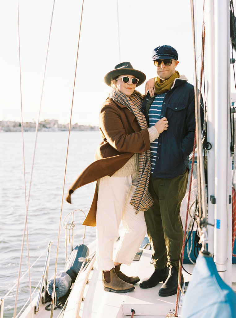 a couple poses for their wedding engagement shoot as they lean into one another while standing on a boat in the ocean near Newport Beach, California. Photographed by wedding photographer Kim Branagan