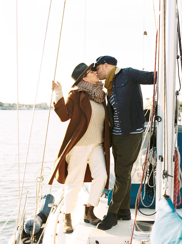 A newly engaged couple kiss each other as they stand on a boat in Newport Beach, California posing for their wedding engagement photos. Photographed by wedding photographer Kim Branagan