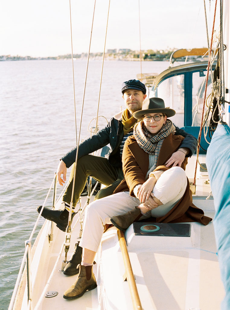 a woman sits towards the front of a sailboat while her fiance sits behind her as they pose, smiling, for their engagement shoot. By wedding photographer Kim Branagan.