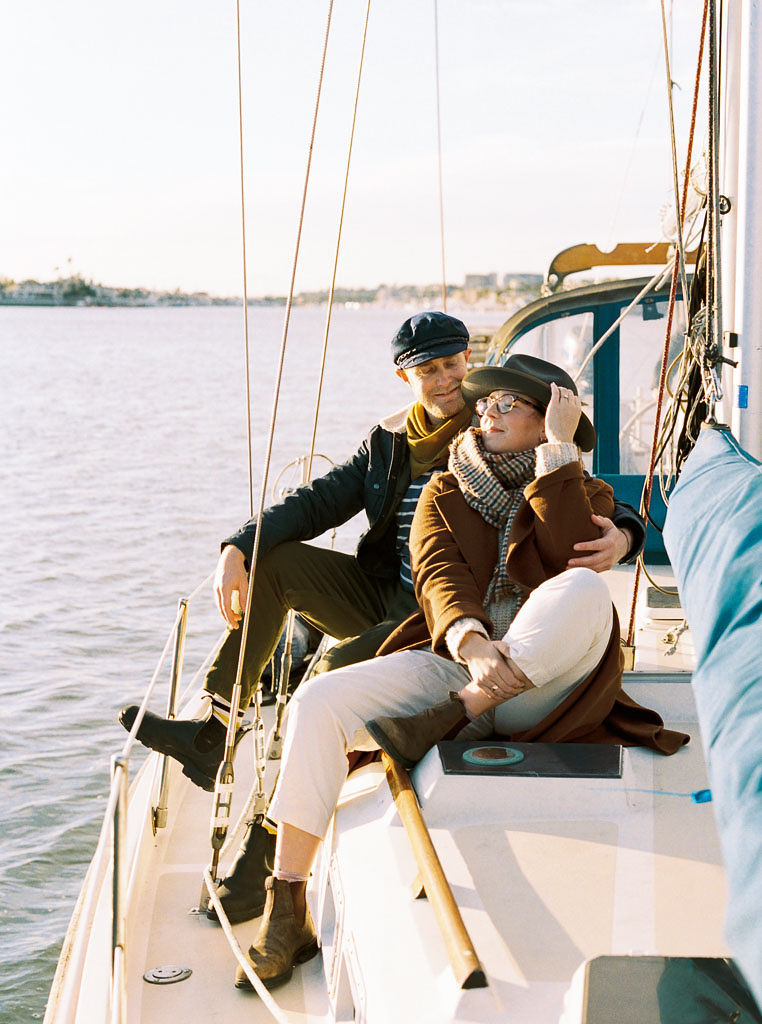 A man and woman sit on a sailboat smiling and embracing during an engagement photo session in Newport California by wedding photographer Kim Branagan