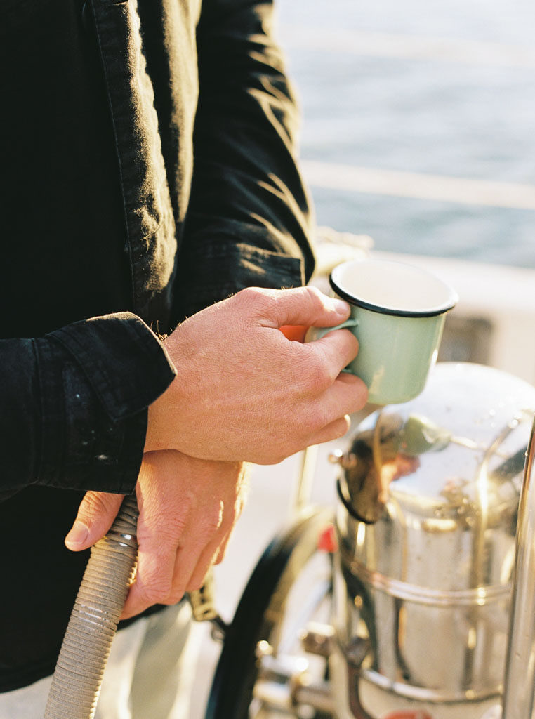 a mans hand holds onto the ships wheel of a sailboat while his other hand holds a tin cup during a wedding engagement photo shoot in Newport Beach, California by wedding photographer Kim Branagan