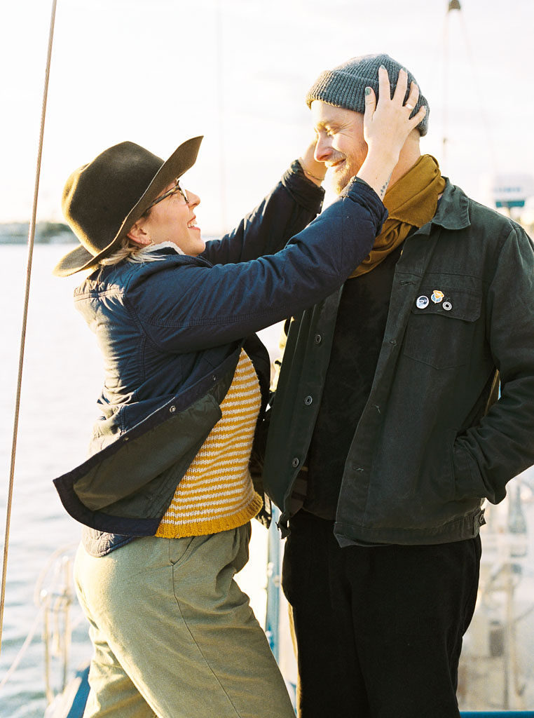 a woman playfully grabs the beanie on her fiances head as she smiles. He stands smiling back at her with his hands in his coat pockets as they pose on a sailboat during their wedding engagement shoot in Newport Beach, California