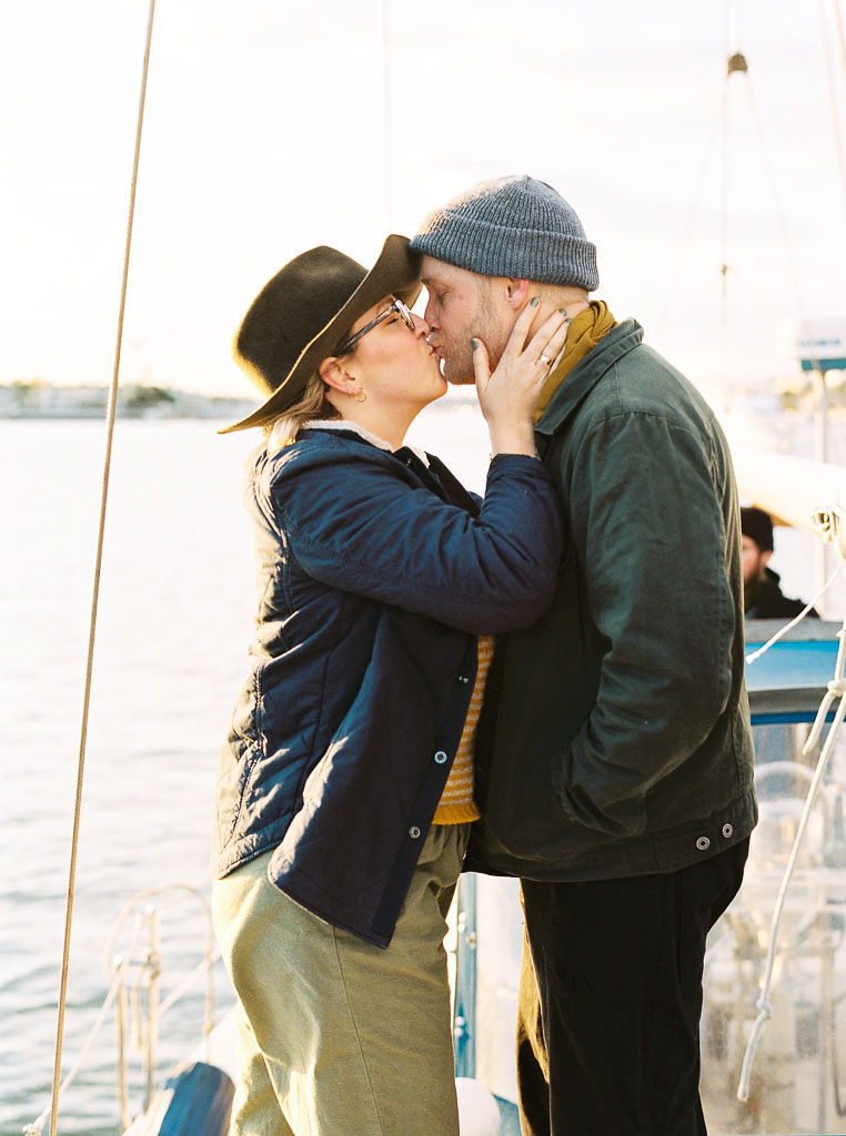 a man and a woman kiss as the woman's hands gently grasp the mans face while his hands are in his coat pockets. They stand on a sailboat in Newport Beach California during their wedding engagement photo shoot.
