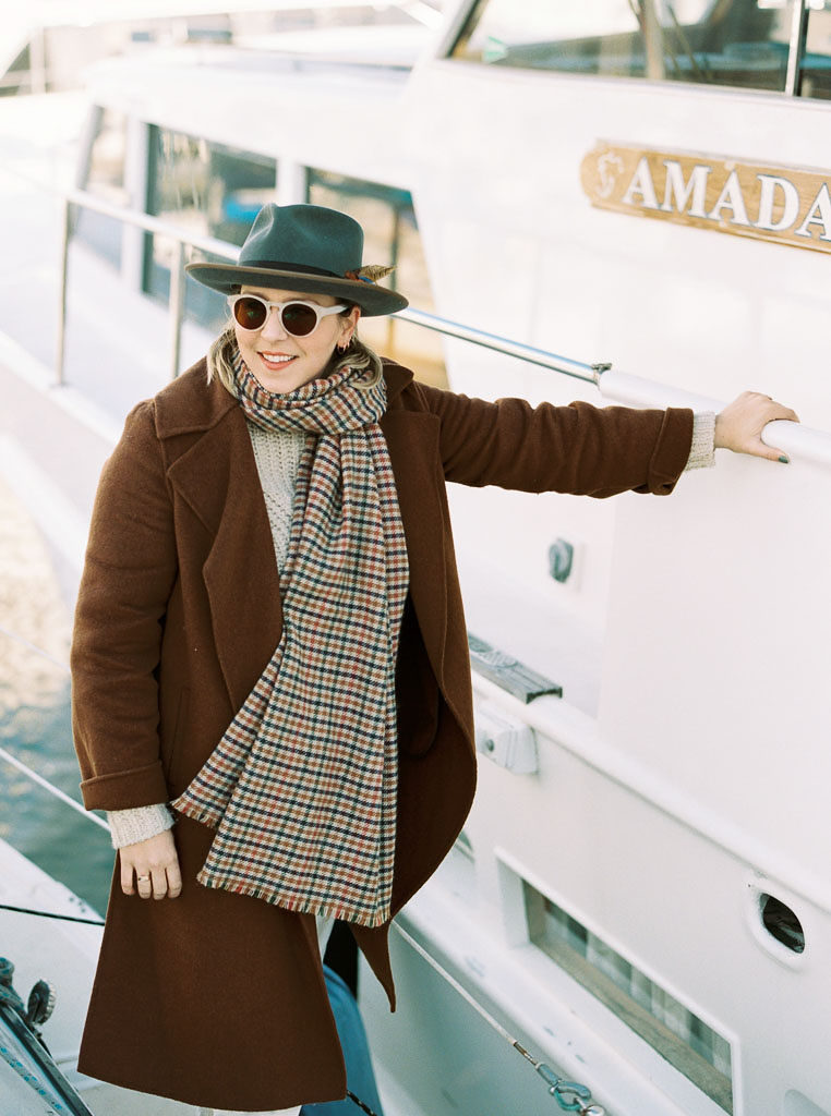 A woman stand next to a boat wearing a scarf, coat, sunglasses and a hat during a wedding engagement shoot in Newport Beach, California by wedding photographer Kim Branagan