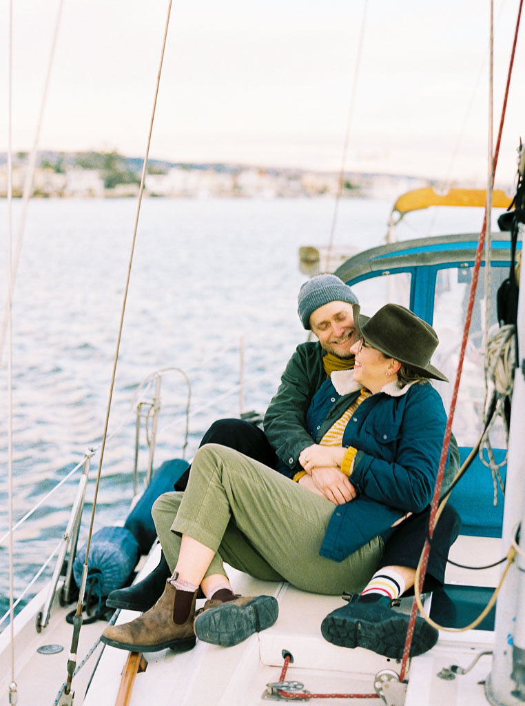 A couple sits towards the front of a sailboat. The woman looks up into her fiance's eyes as he looks down into hers as they both smile during a wedding engagement photo shoot in Newport Beach, California.
