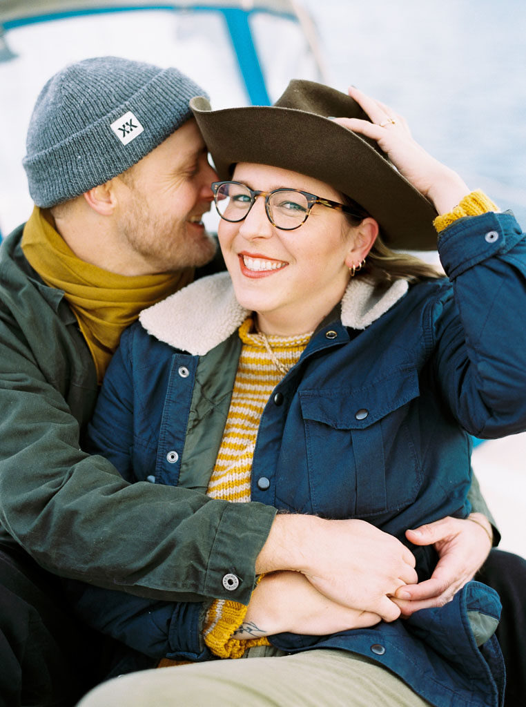 A woman looks at the camera smiling as her fiances wraps his arms around her with his face near her cheek. She holds her Goorin Bros hat with one hand and embraces her fiance's hand with the other during their engagement photo shoot in Newport Beach, California.
