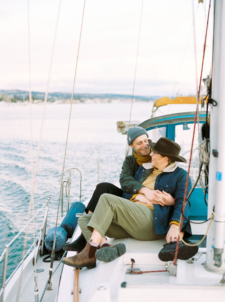 A couple sits on the side of a sailboat embracing and smiling at each other during a wedding engagement photo shoot in Newport Beach, California by wedding photographer Kim Branagan
