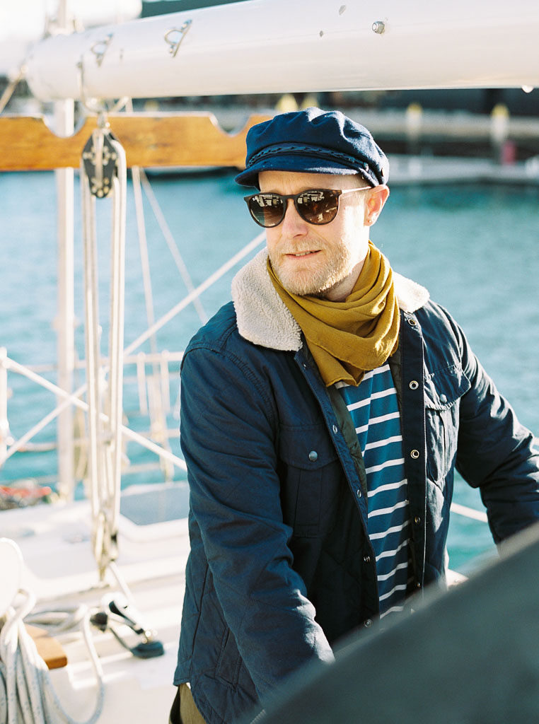 A man wearing a hat and sunglasses poses on a boat while looking into the distance during a wedding engagement shoot in newport, CA. By Newport wedding photographer Kim Branagan