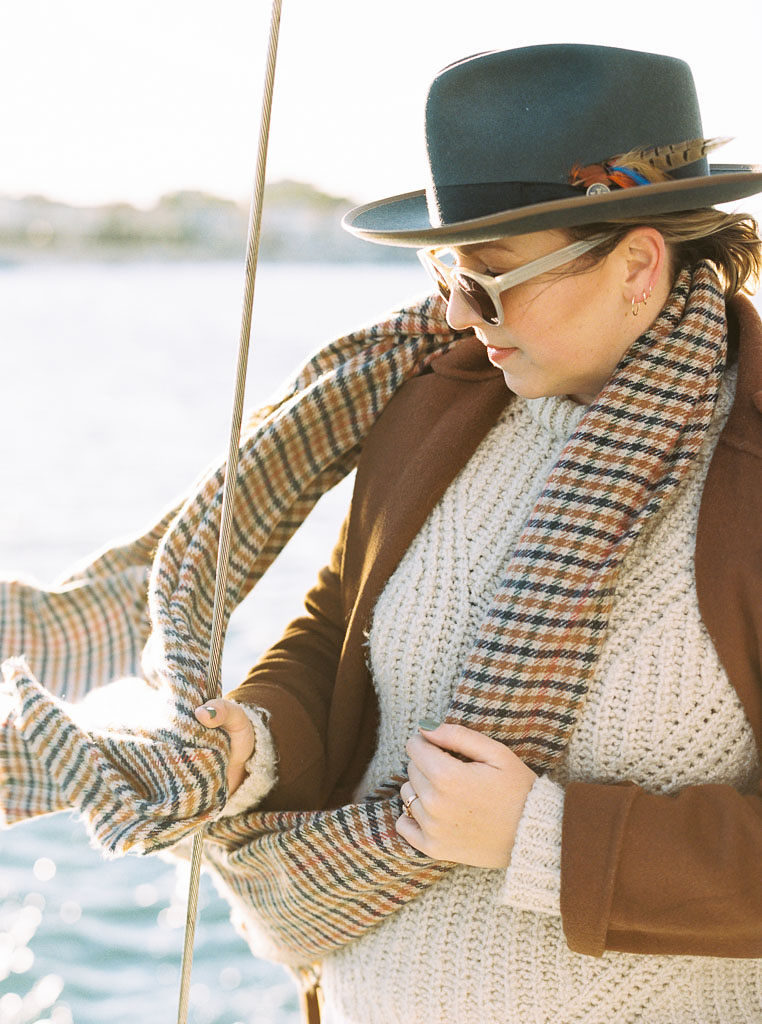 A woman with a hat on and sunglasses stands with the pacific ocean behind her as her scarf blows in the wind during engagement shoot in Newport Beach by DC wedding photographer Kim Branagan