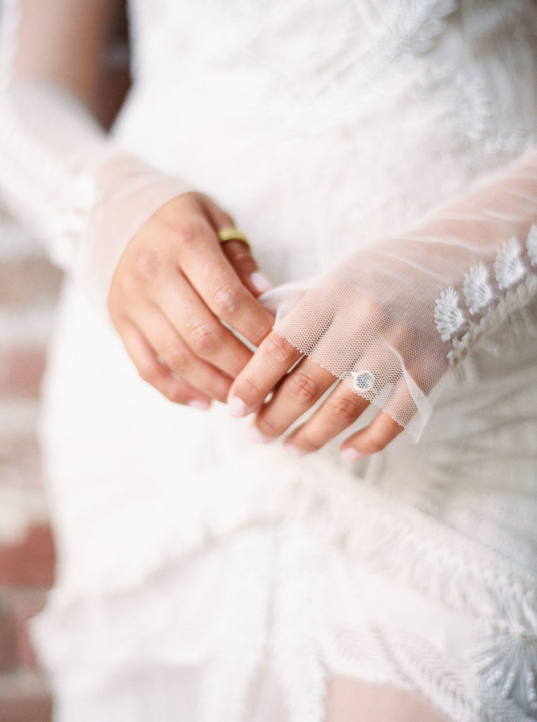 Close-up of bride's hands with gold ring on right thumb and wedding ring on left hand and long sheer sleeves on white wedding gown by photographer Kim Branagan