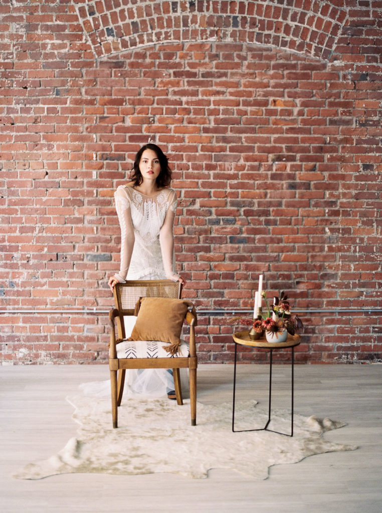 Dark-haired bride wearing white dress and leaning forward on tan chair beside tan stand holding flower bouquets on top of white fur rug and in front of brick wall at a Minimalist Bridal Editorial Shoot