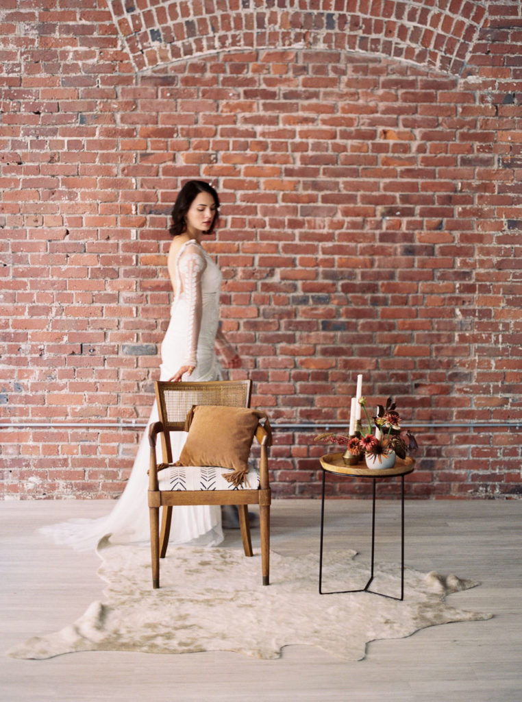 Side view of woman wearing white bridal gown with train and resting her hand on tan wooden chair with brick wall backdrop, tan table holding flowers, and white fur throw rug