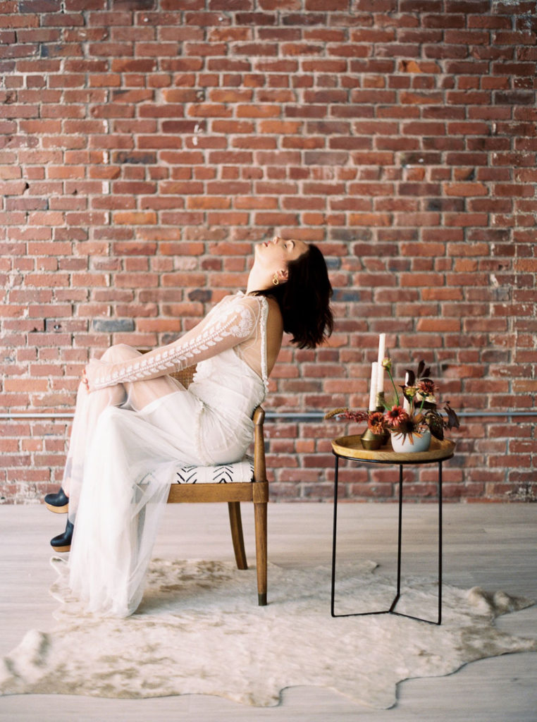 Bride in white gown sitting sideways on wooden chair in front of brick wall with face turned up and dark hair hanging back by Kim Branagan Photography