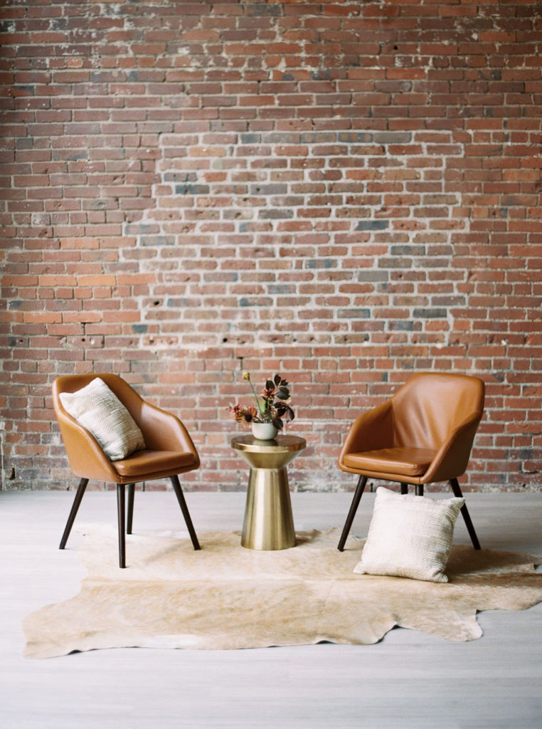 Two tan barrel chairs angled toward each other with gold round stand between them and a light tan fur rug on the floor in front of brick wall by DC wedding photographer Kim Branagan