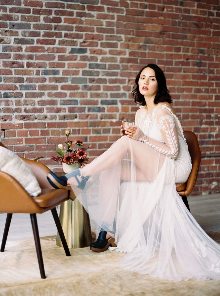 Dark-haired bride wearing sheer and lace wedding gown and black clogs and sitting on tan barrel chair with one foot propped on other tan barrel chair in this Minimalist Bridal Editorial Shoot at The 101 in Seattle, Washington by destination photographer Kim Branagan