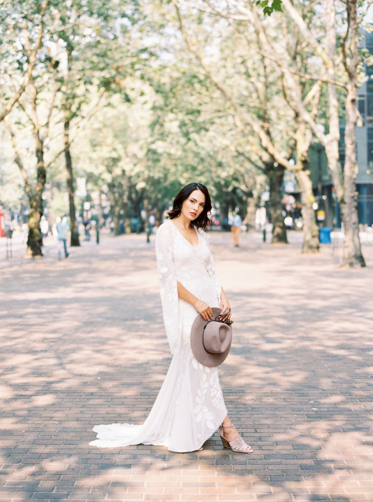 Full-length shot of dark-haired bride wearing white wedding gown and holding gray hat on brick tree-lined promenade