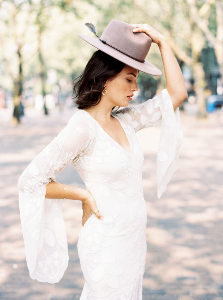 Dark-haired woman holding up gray hat and looking down while wearing white bridal gown with flared sleeves by VA photographer Kim Branagan