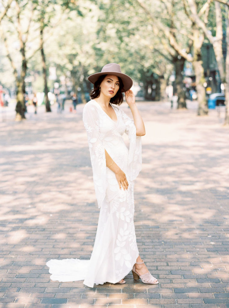 Full-length photo of bride in gray rimmed hat and white wedding dress with train and soft gray wedding shoes on brick promenade