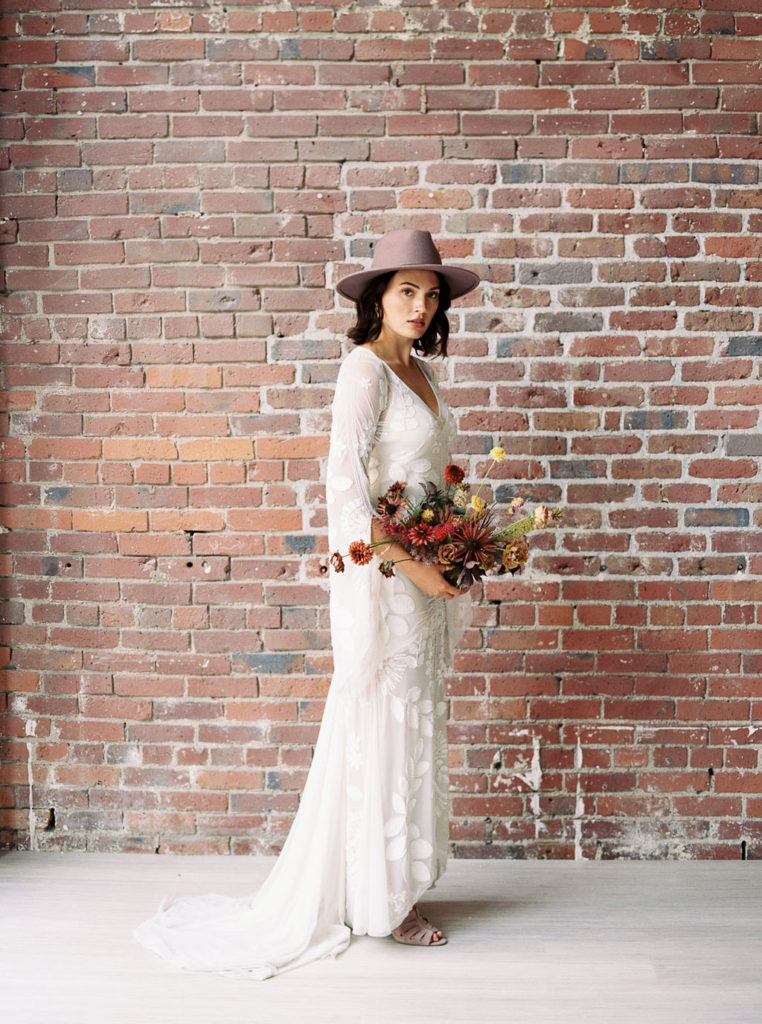 Full-length shot of bride in white wedding gown with train and lavendar fedora and holding fall bouquet in front of brick wall in Minimalist Bridal Editorial Shoot at The 101 in Seattle, Washington by destination photographer Kim Branagan