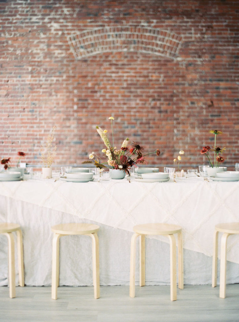 Bridal table setting with white tablecloth, white dishware, light tan chair stools, and fall-colored flower bouquets by wedding photographer Kim Branagan