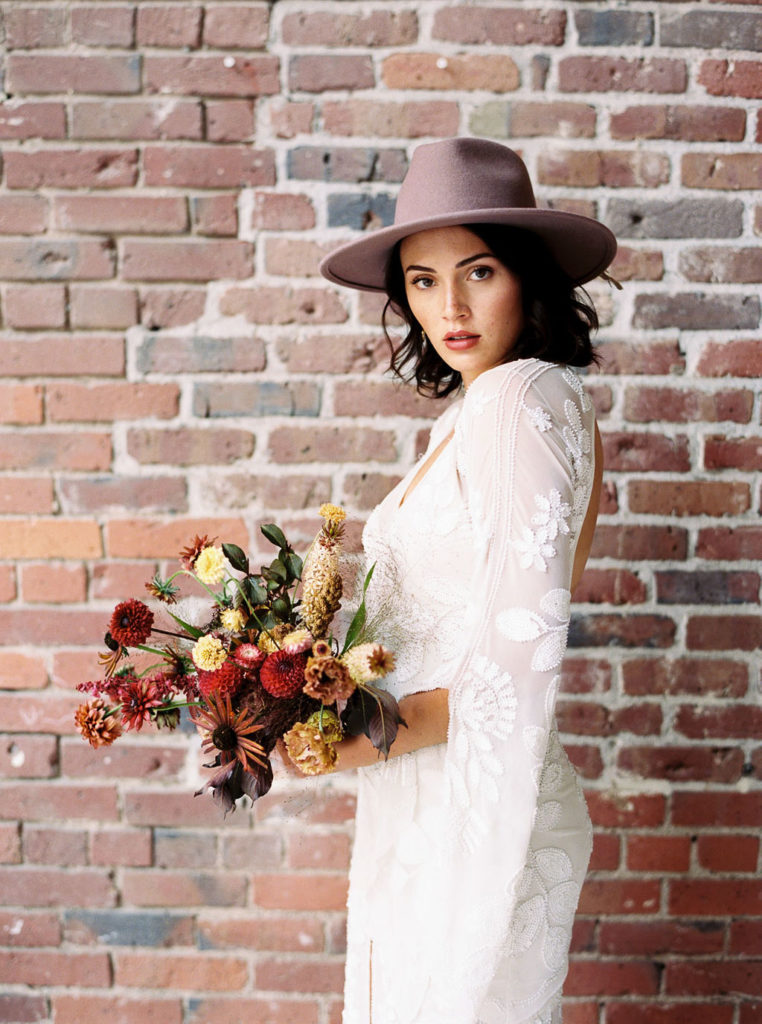 Side shot of dark-haired woman in lavender fedora and white bridal gown in front of brick wall and holding bouquet of fall flowers in Minimalist Style Bridal Photoshoot
