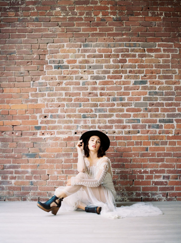 Bride wearing white wedding gown, black clogs, and black fedora hat and sitting on white floor in front of brick wall by Kim Branagan Photography