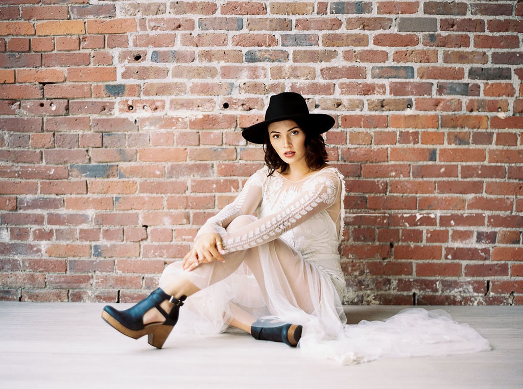 Bride wearing sheer and lace wedding gown, black hat, and black clogs with brick wall backdrop by wedding photographer Kim Branagan
