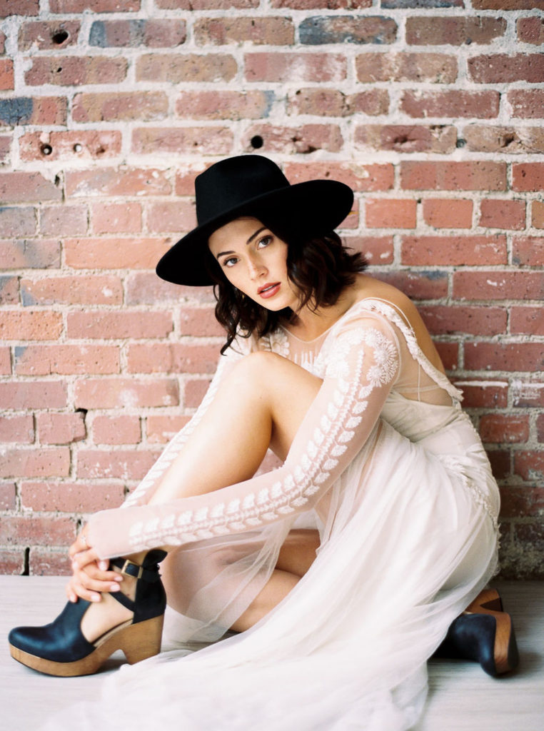 Bride wearing long-sleeved lace white wedding gown, black fedora, and black strappy clogs and sitting cross-legged in front of brick wall