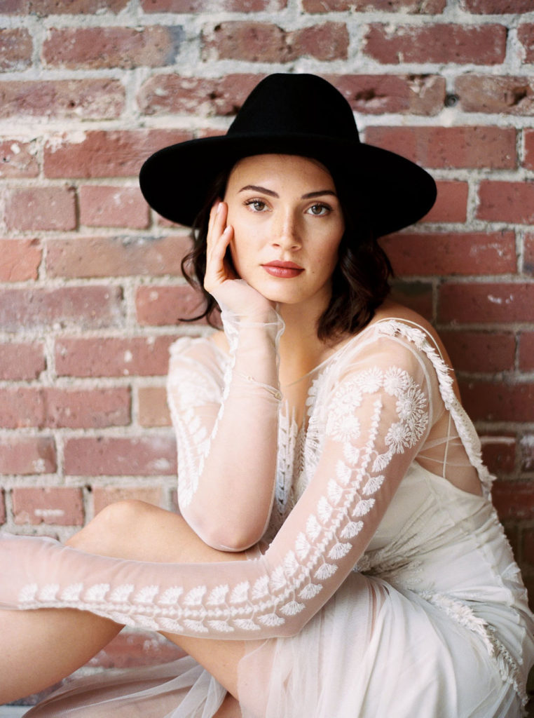Woman wearing black brimmed hat and white dress with sheer long sleeves and sitting in front of brick wall while looking at the camera by DC photographer Kim Branagan