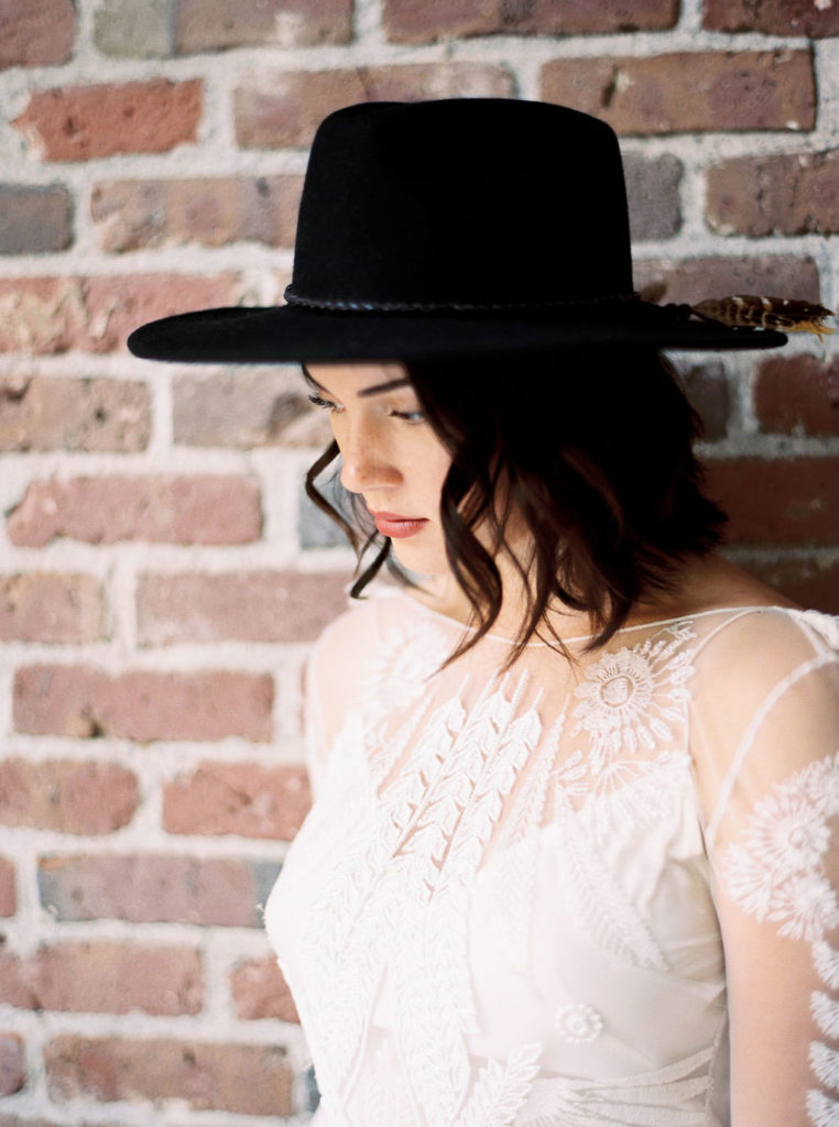 Woman wearing black fedora hat accented with feathers and a white lace bridal dress, standing in front of brick wall, and looking down