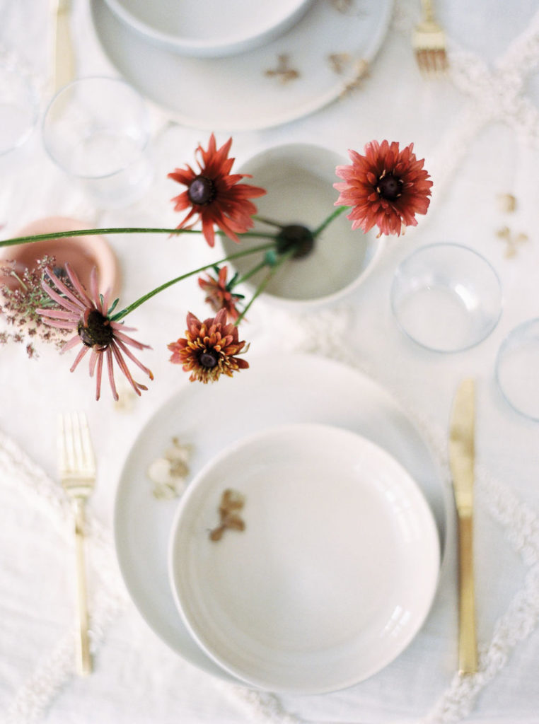 Overhead shot of white tableware with gold accents and gold cutlery accented by long-stemmed mauve and rust-colored flowers by wedding photographer Kim Branagan