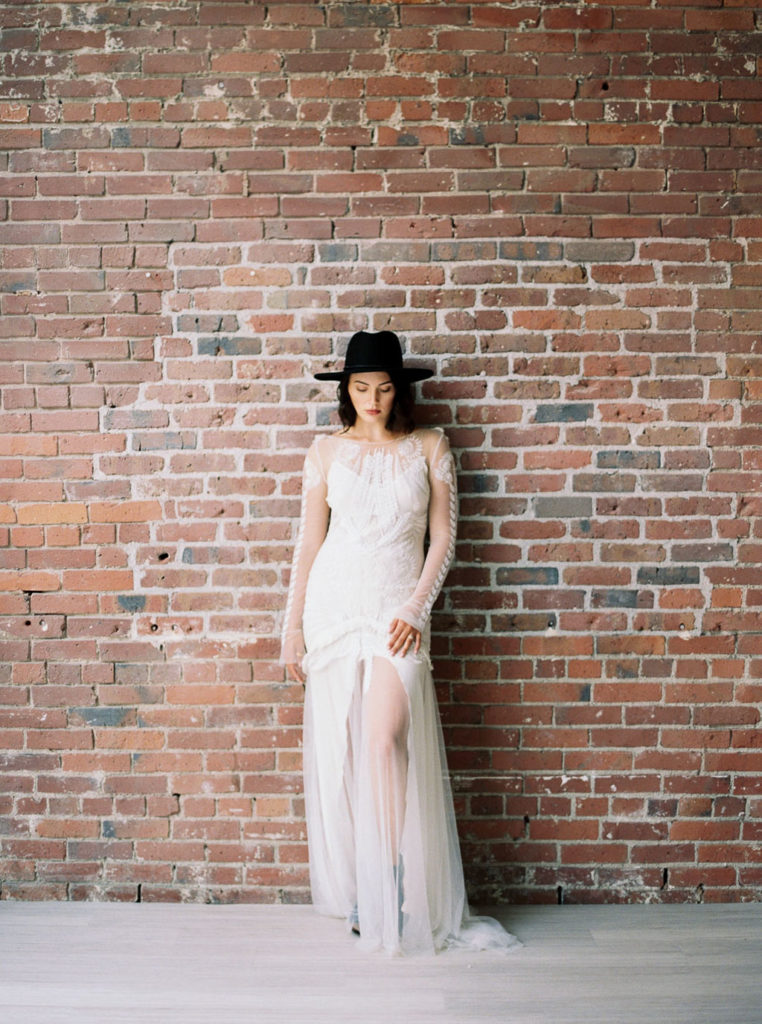 Full-length shot of bride wearing long-sleeved white wedding dress and black hat and standing in front of brick wall in Minimalist Bridal Editorial Shoot at The 101 in Seattle, Washington by destination photographer Kim Branagan