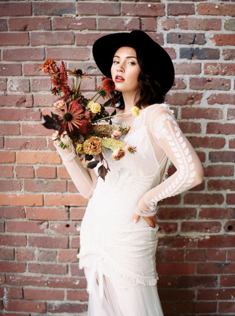 Bride holding fall bouquet of yellow, mauve, and rust-colored flowers and wearing black brimmed hat and white long-sleeved gown at a Minimalist Bridal Editorial Shoot by Kim Branagan Photography