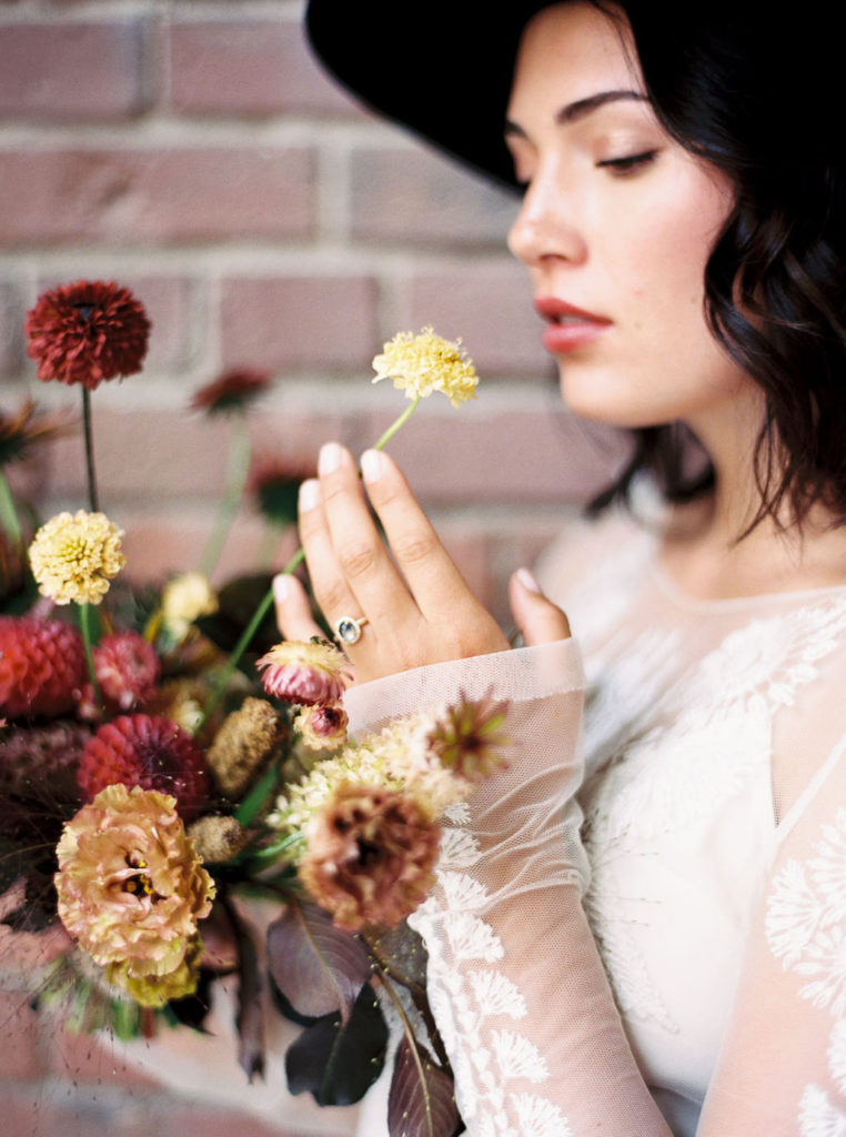 Side view of woman holding bouquet of fall flowers and holding yellow dress up toward her nose and wearing black rimmed hat and white long-sleeved dress and wedding ring