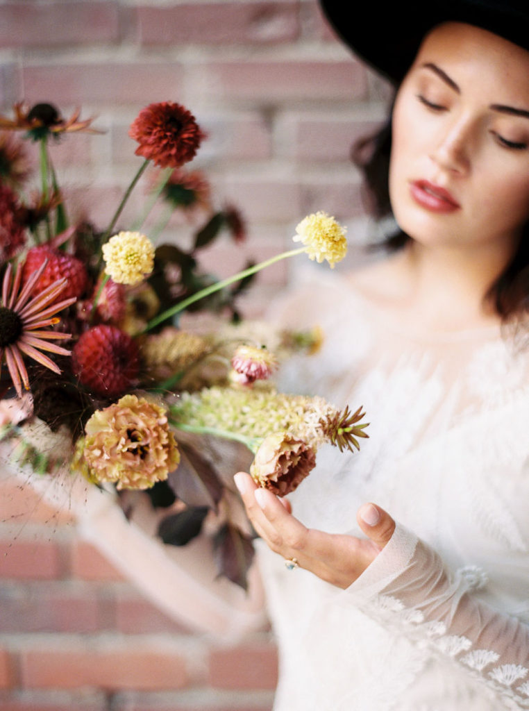 Bride wearing long-sleeved white dress and looking down to examine fall-colored flower bouquet that she is holding