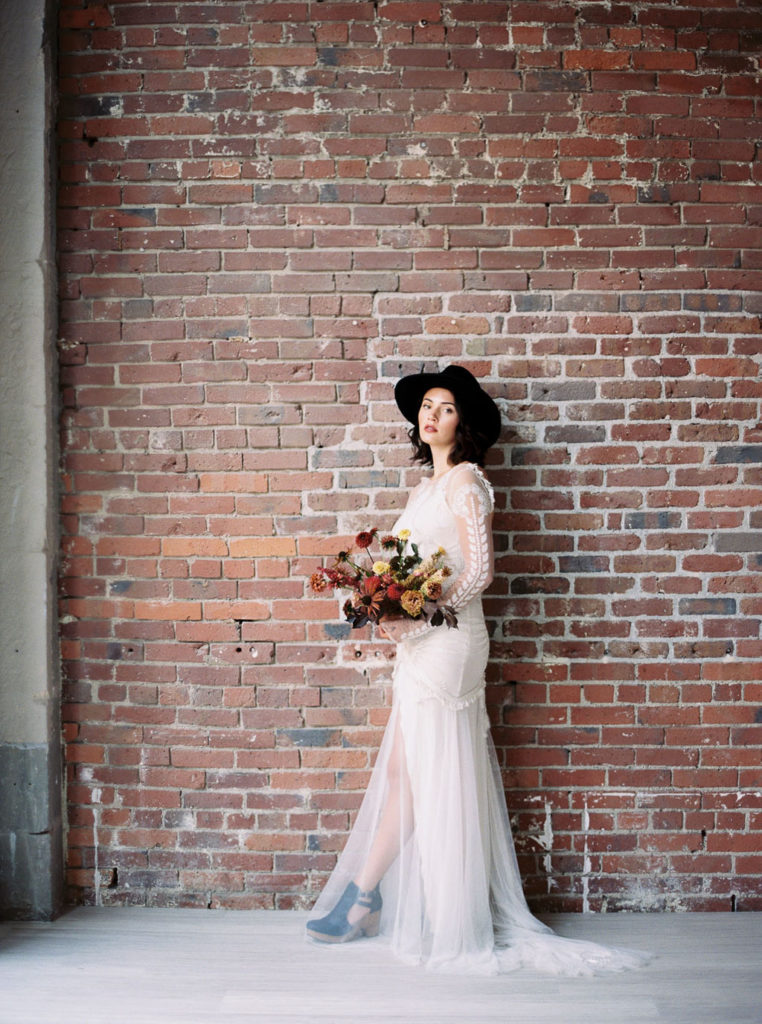 Full-length shot of woman in white wedding gown and black rimmed hat and wearing black shoes while holding fall-colored flower bouquet at a Minimalist Bridal Editorial Shoot
