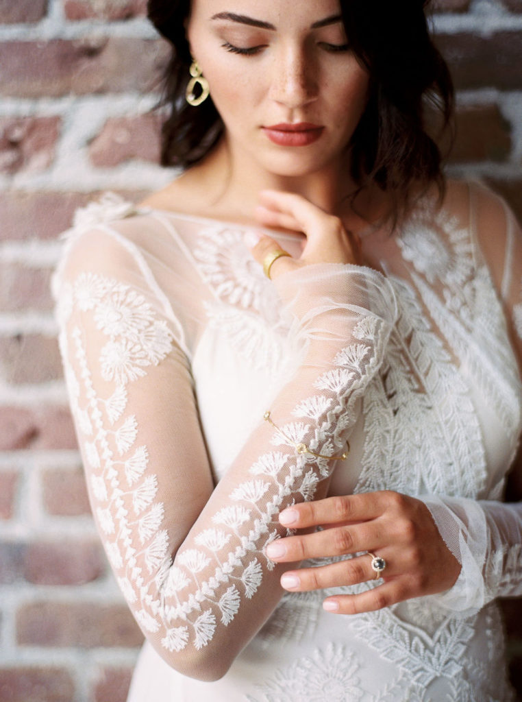 Woman wearing small gold hoop earrings, long-sleeved lace appliqued wedding gown, and wedding ring and looking down in Minimalist Style Bridal Photoshoot