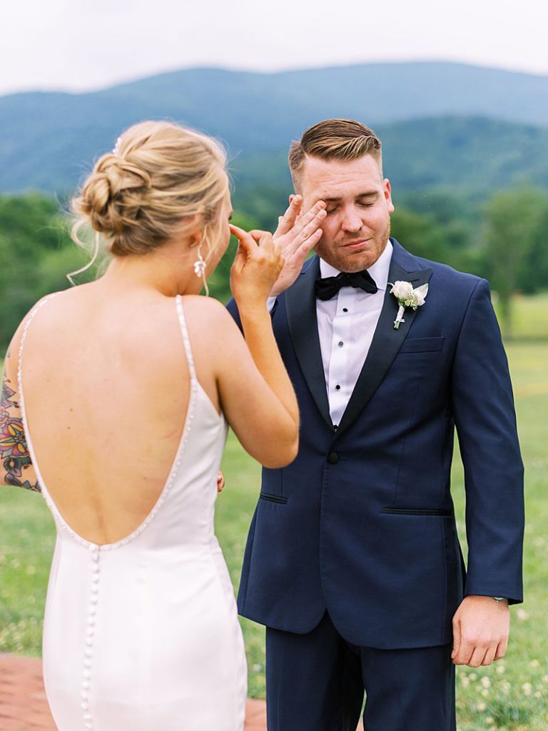 A groom stands in front of his bride as he wipes a tear from his eye