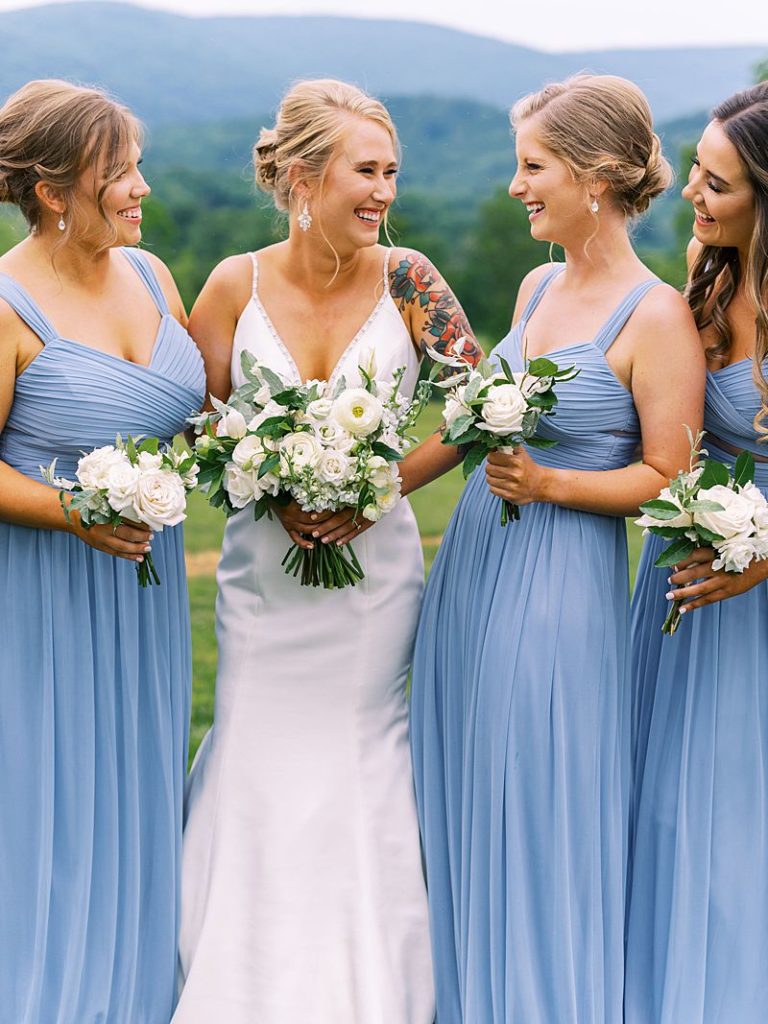 A bride holds a white bridal bouquet while smiling at her bridesmaids as they stand on either side of her smiling back