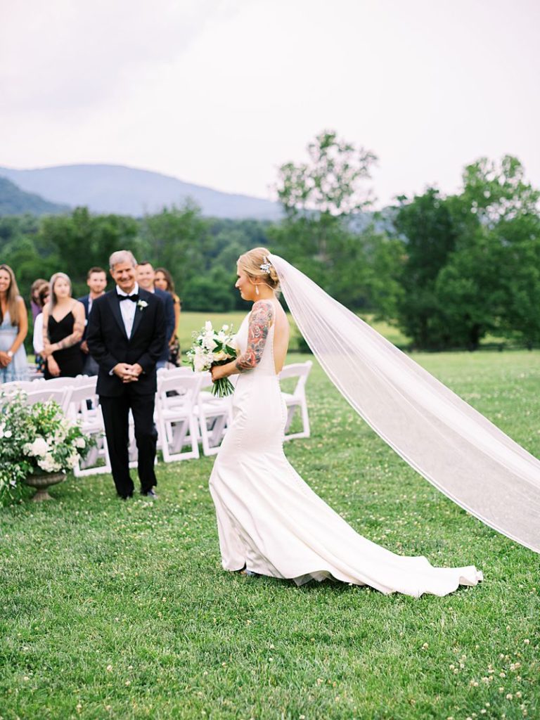 A bride walks down the aisle at her wedding outside at King Family Vineyards. Her white vail flows behind her in the wind. Photographed by Virginia wedding photographer Kim Branagan