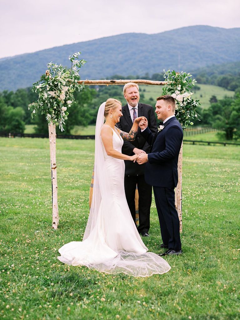 A bride and groom stand at the alter outside with a minister and a floral archway in the background at King Family Vineyards in Virginia.