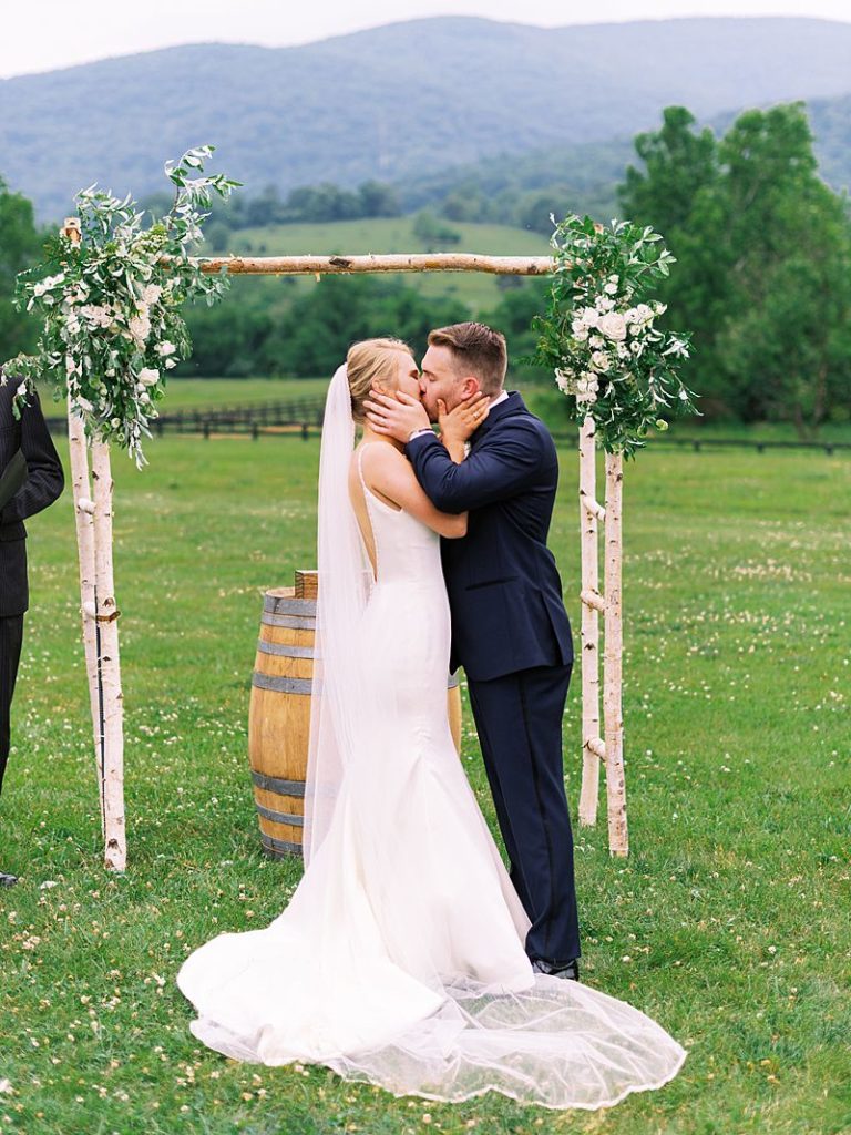 a bride a groom kiss at the altar with a floral arch behind them while standing outside at King Family Vineyards