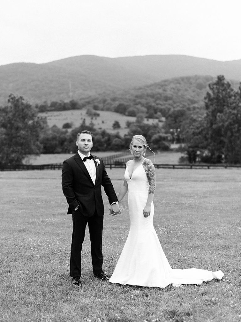 bride and groom hold hands and look, straight-faced, at the camera while standing outside at King Family Vineyards during a Virginia wedding.
