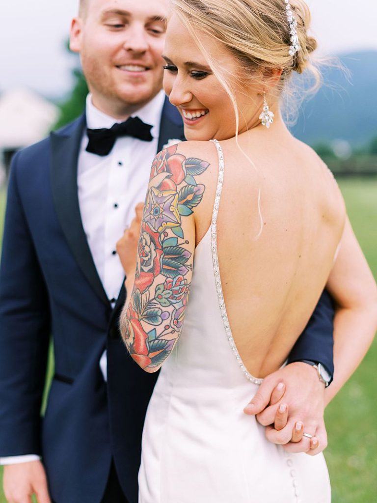 A bride has her back turned to the camera while looking back over her shoulder smiling. Her groom has his hand in hers as they smile.