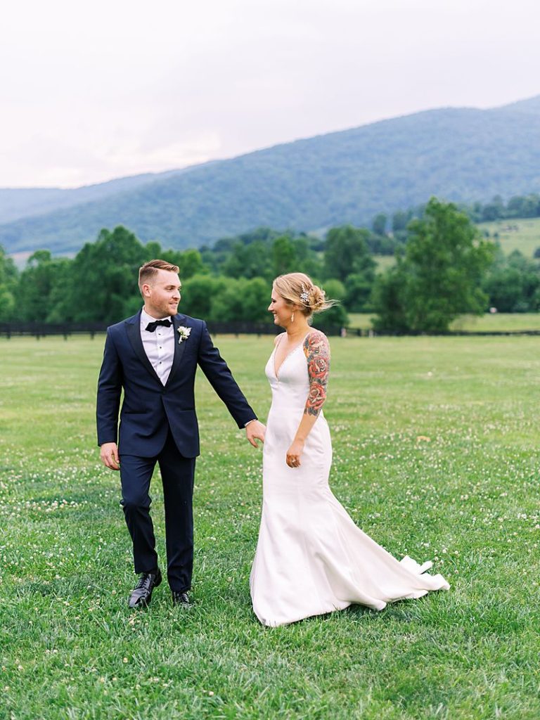 bride and groom walk across a green lawn holding hands smiling. Tall, tree covered hills are in the background at King Family Vineyards in Virginia.