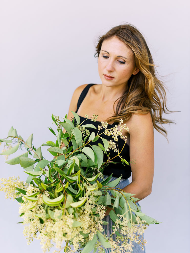 Floral designer with sandy-colored hair holding bouquet of greenery and cream flowers by portrait photographer Kim Branagan