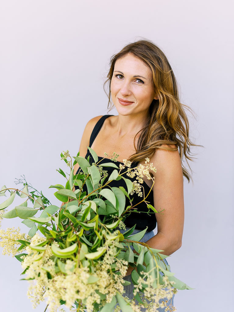 Smiling woman with sandy-colored hair holding bouquet of greenery and cream flowers by branding photographer Kim Branagan