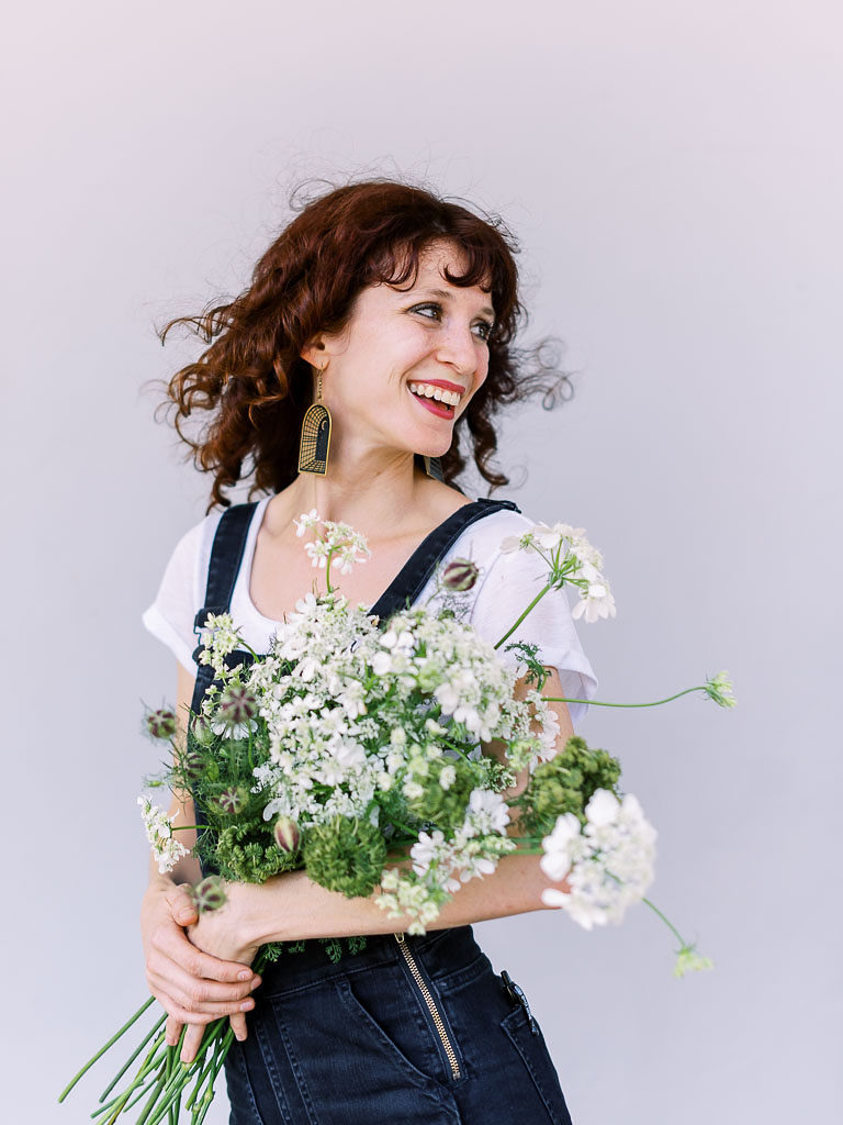 Chestnut-haired woman holding bouquet of greenery and white flowers and looking back and smiling by branding photographer Kim Branagan