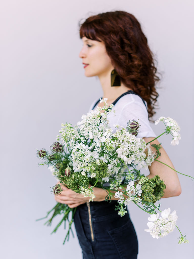 Side portrait shot of chestnut-haired woman in white top and black pants holding bouquet of fresh white flowers and green accents